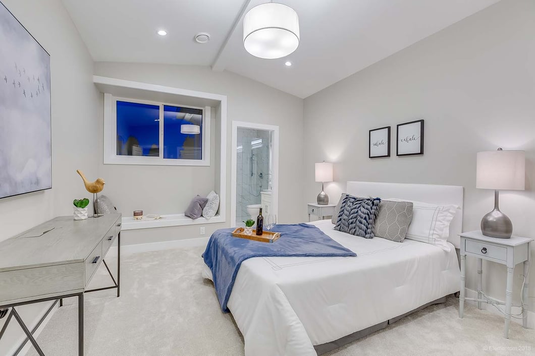 Bright modern bedroom with neutral décor, white bedding, blue accents, bedside lamps, recessed lighting, and a window seat.
