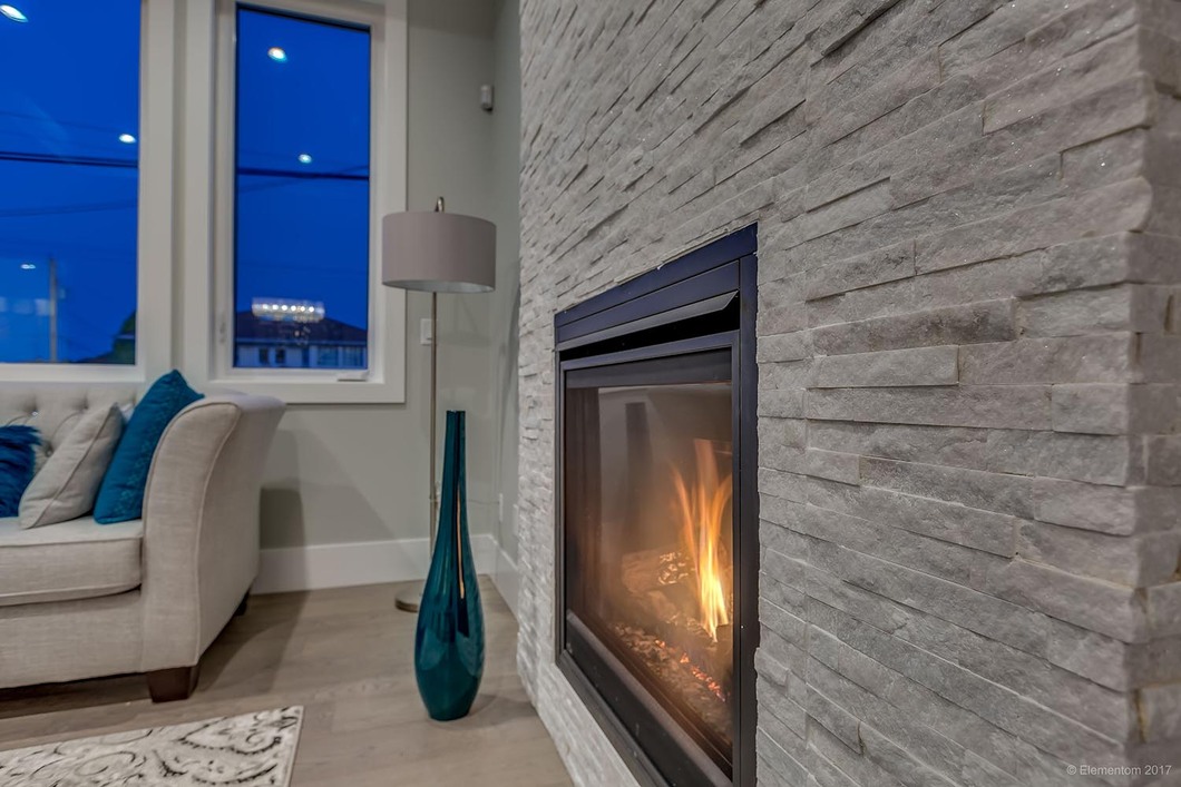 Modern fireplace with stacked stone surround, warm flames, blue accent décor, and large windows beside a neutral sofa.