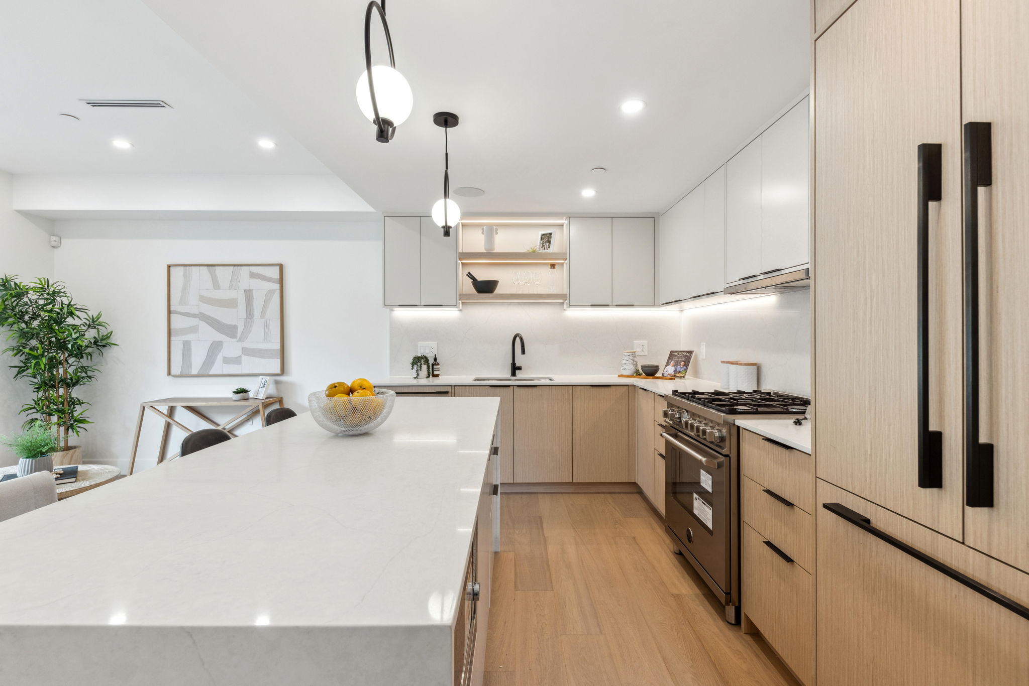 Modern kitchen with light wood cabinetry, white quartz island, black fixtures, pendant lighting, and warm wood floors.