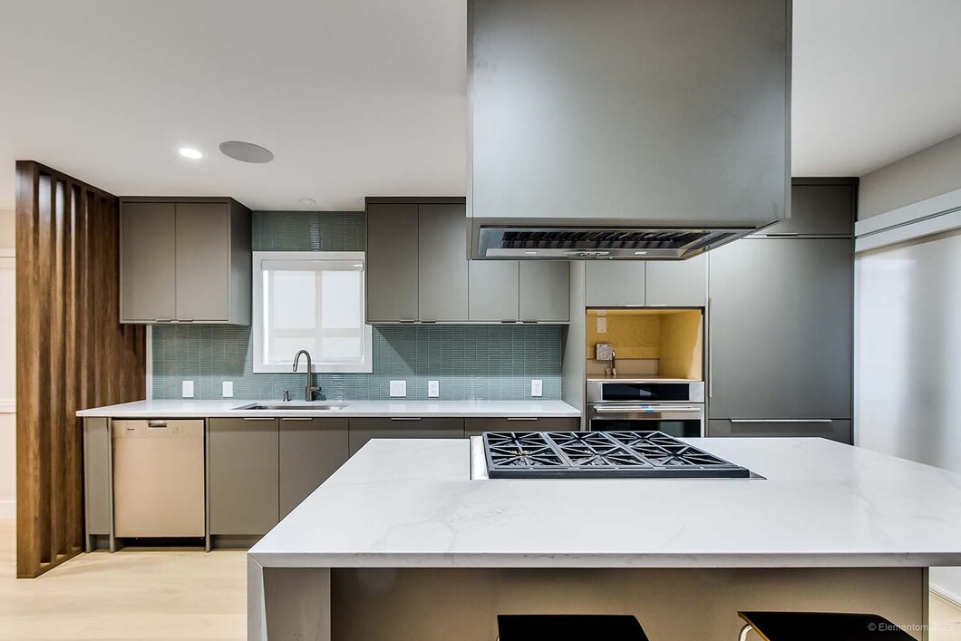 Modern kitchen with quartz island, gas cooktop, suspended range hood, grey cabinetry, wood accents, and tile backsplash.